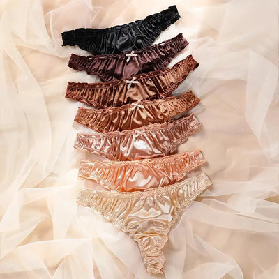 Top-down flat lay of seven ruffled satin thong panties arranged by color on sheer white fabric