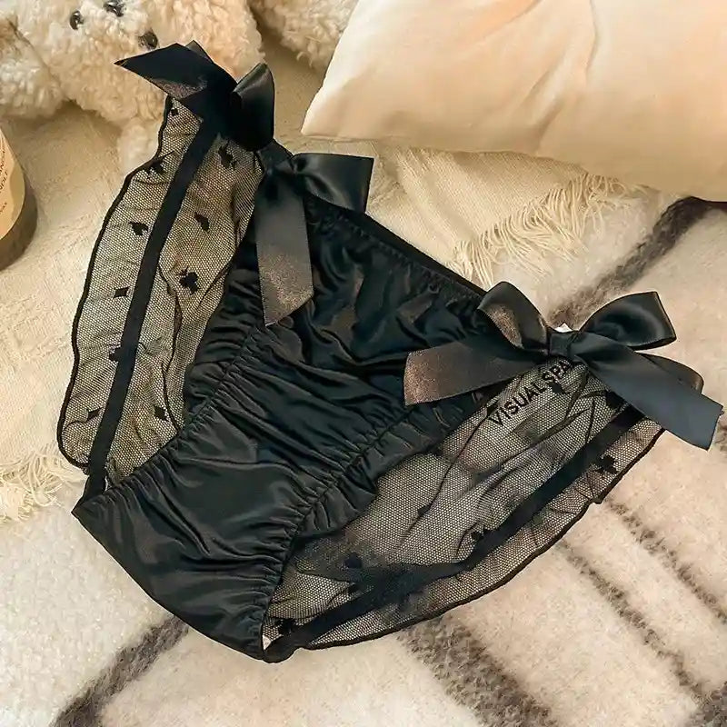 Black satin and mesh panties with ribbon bow ties on each hip and embroidered sheer side panels, flat lay on textured bedding