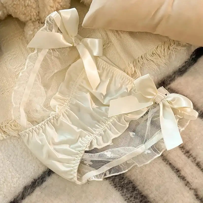 Ivory bloomer panties with satin ribbon bows, lace trim, and sheer mesh panel, laid flat on a woven blanket